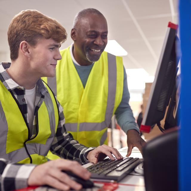 Intern With Supervisor Working In Busy Modern Warehouse On Computer